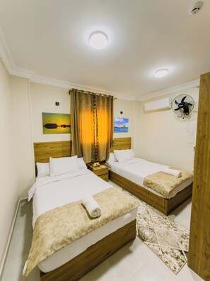 Une chambre d’hôtel avec deux lits, une table de chevet, un ventilateur fixé au mur et un tableau représentant un coucher de soleil.