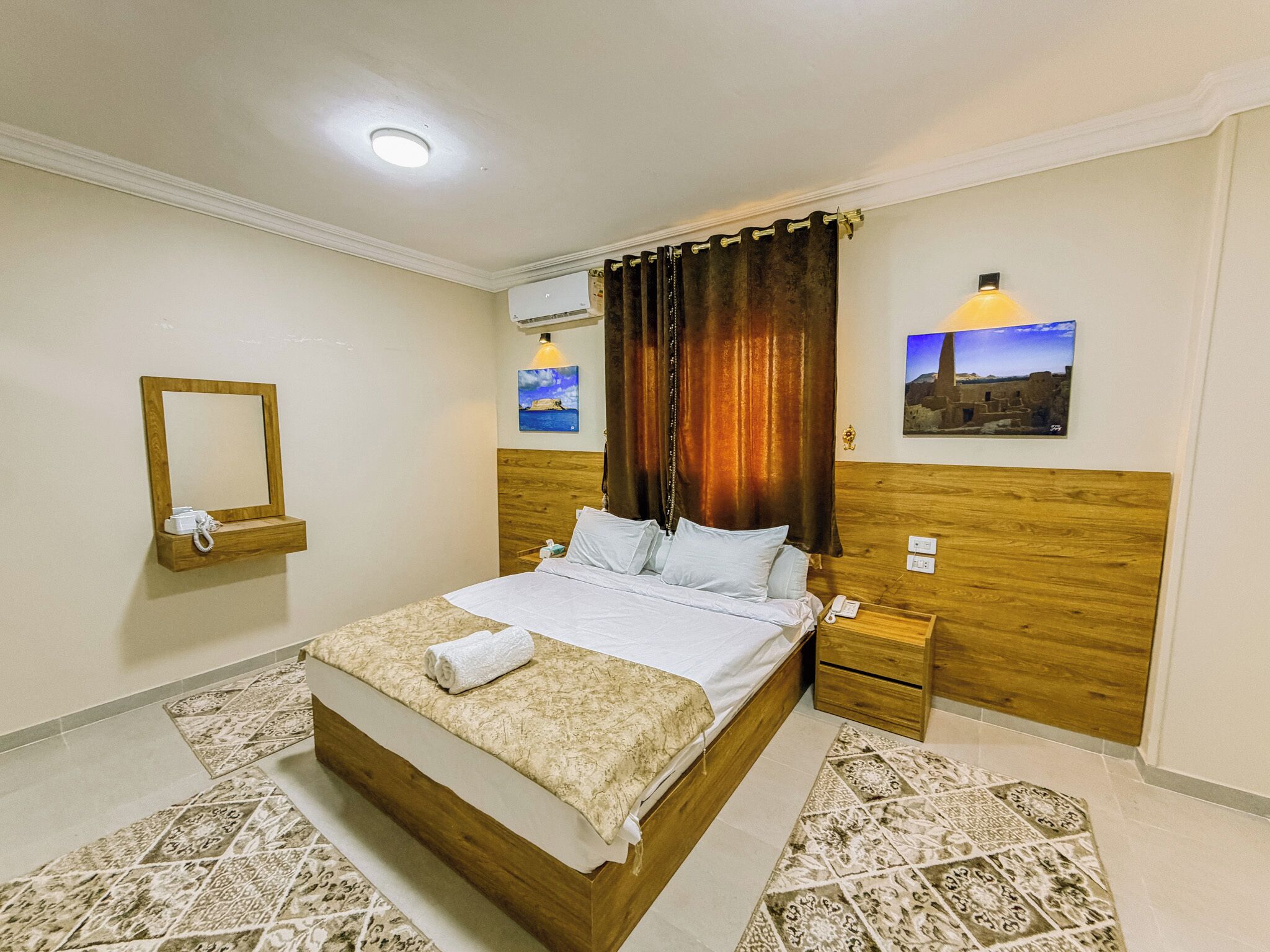Una habitación de hotel con cama, mesitas de noche, un espejo, un equipo de aire acondicionado y obras de arte en las paredes.