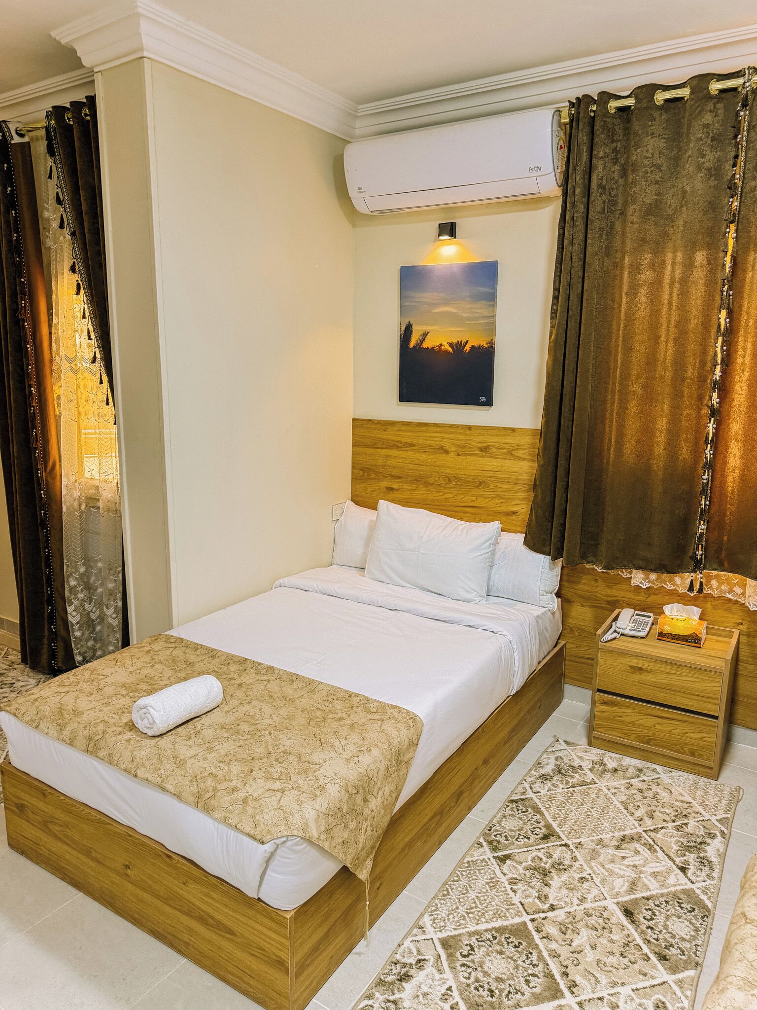 Habitación de hotel con cama, mesita de noche, equipo de aire acondicionado, un cuadro y cortinas.
