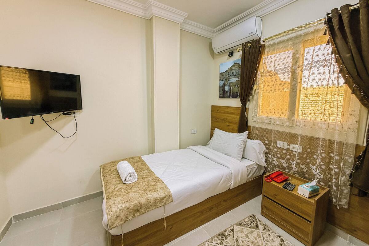 Une chambre à coucher comprenant un lit, un téléviseur fixé au mur, un appareil de climatisation, une fenêtre avec des rideaux et une table de chevet avec un téléphone et des mouchoirs.