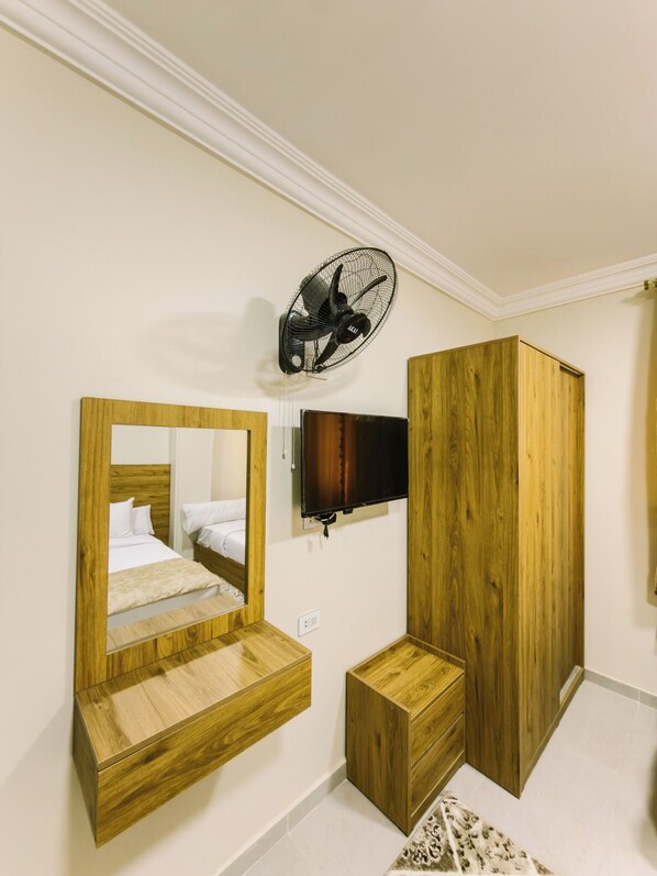 A bedroom with a bed, a wooden wardrobe, a TV, a mirror, and a fan.