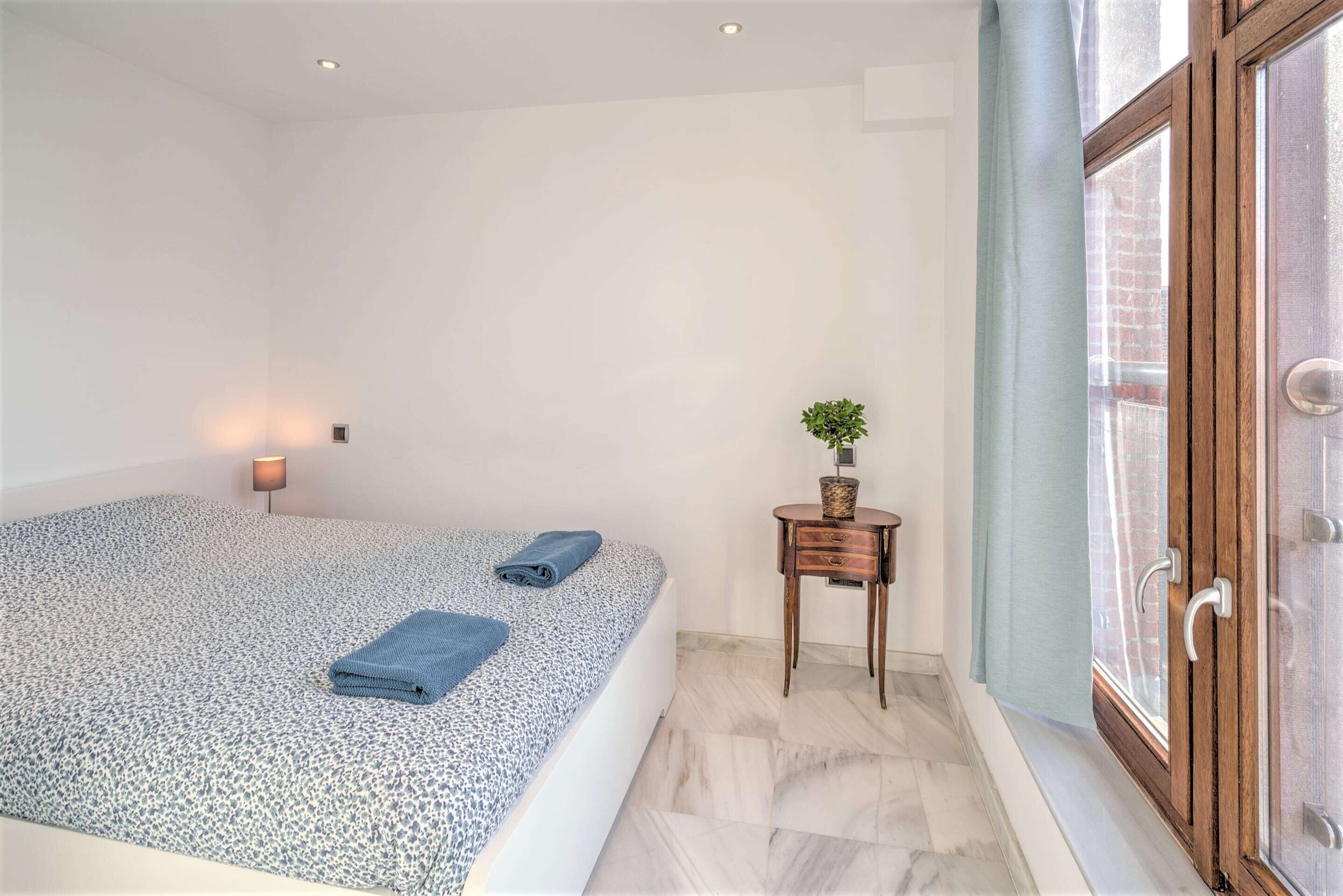 Een slaapkamer met een bed, nachtkastje met lamp, raam met gordijnen en een deur.