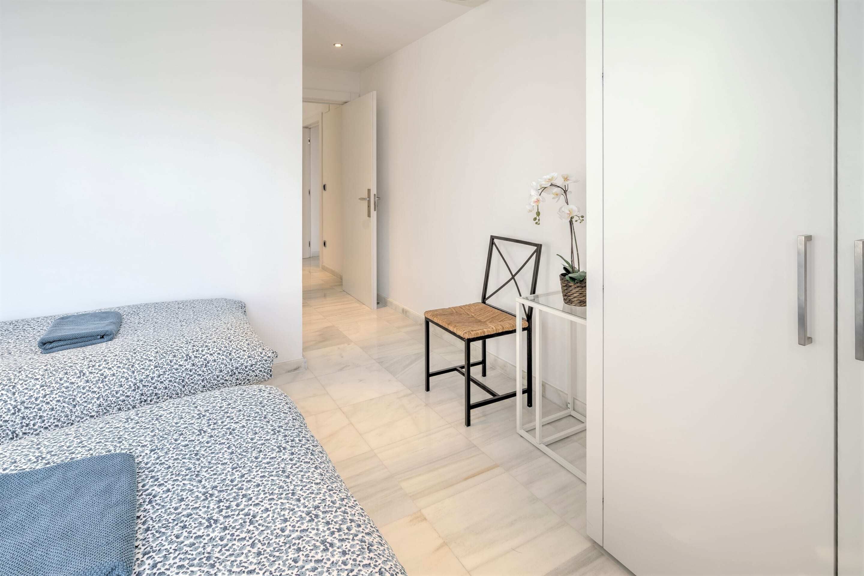 A modern bedroom with two beds, a chair, a small table with a plant, and a white wardrobe.