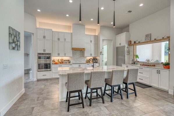 A modern kitchen with white cabinetry, a central island with bar stools, a built-in oven, and pendant lighting. - Amazing 4-bedroom House with Pool, Golf Simulator, and Batting Cage. (Lago Vista)