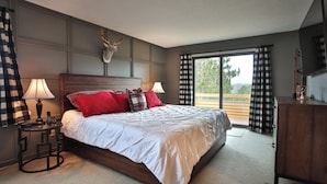 Une chambre avec un grand lit, deux tables de chevet avec des lampes, une tête de lit en bois, une tête de cerf fixée au mur et une vue sur l’extérieur, accessible par une porte-fenêtre coulissante.