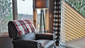Une pièce avec un fauteuil en cuir, un lampadaire, un rideau à carreaux et une vue par la fenêtre.