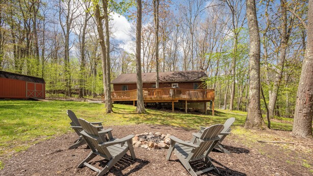 A cabin with a deck, surrounded by trees, and a fire pit with chairs. - New! Lake Area Chalet w/ Hot Tub (Swanton)