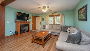 A living room with a fireplace, a ceiling fan, a television, and a couch. - New! Lake Area Chalet w/ Hot Tub (Swanton)