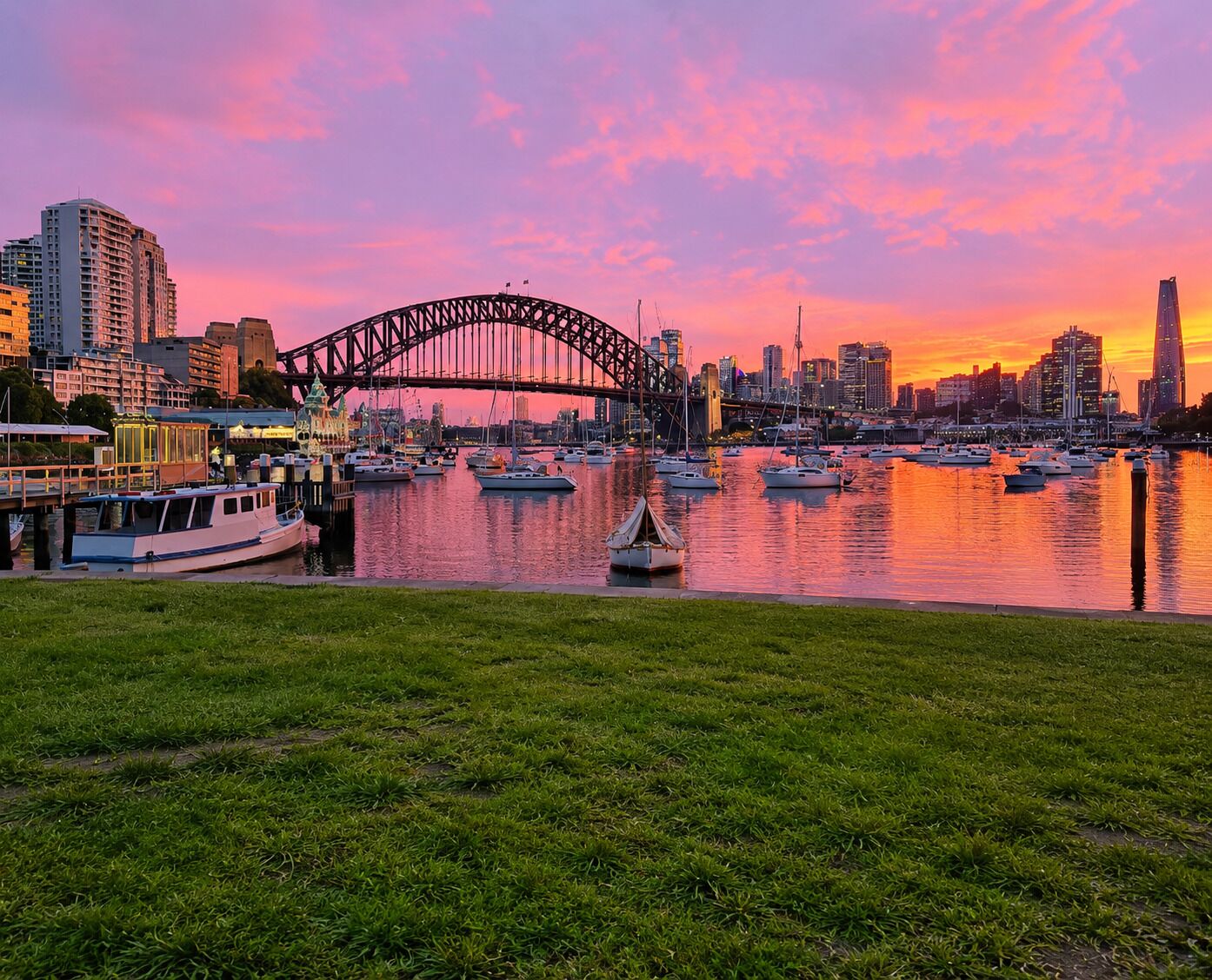 A cityscape with a prominent bridge, boats on the water, and a grassy foreground.