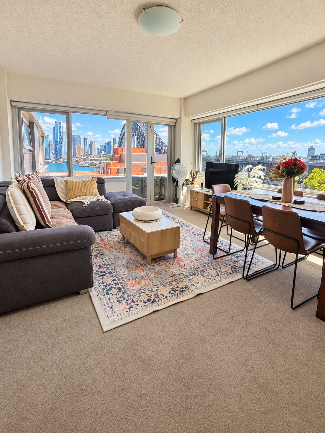 A modern living room with a sofa, coffee table, and dining area with chairs. Large windows offer a view of the city skyline.