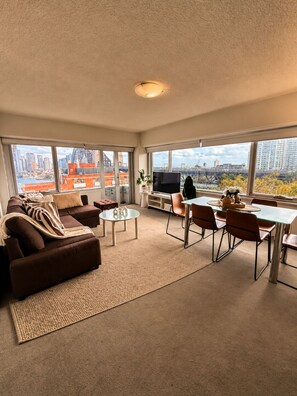 A modern living room with a sofa, coffee table, and dining area, large windows with a city view. - Iconic Harbour Views & Free Parking (Kirribilli)