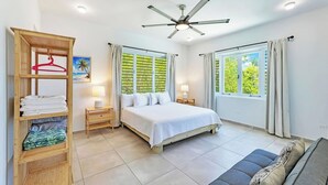 Ein Schlafzimmer mit Bett, Nachttischen, einem Kleiderschrank, einem Deckenventilator und großen Fenstern mit Vorhängen.