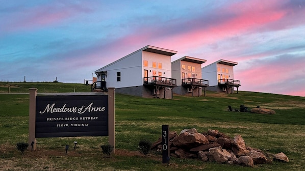 A private retreat with three buildings, a grassy hill, and a sign indicating the name and location. - Cozy Modern Retreat in Floyd with Fire Pit and Peaceful Mountain Views (Floyd)