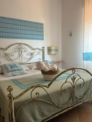 Una cama con estructura metálica, una cesta tejida sobre la cama y una lámpara fijada a la pared.