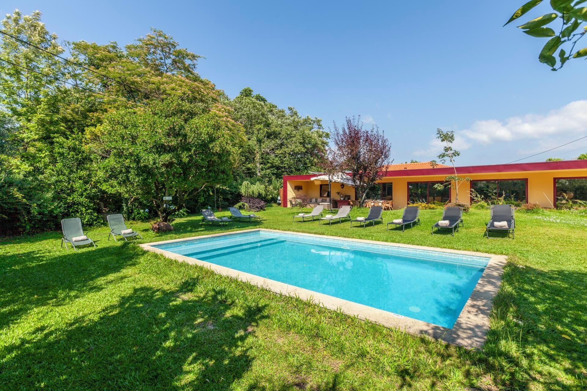 A swimming pool with lounge chairs, surrounded by greenery and a building with a red roof.