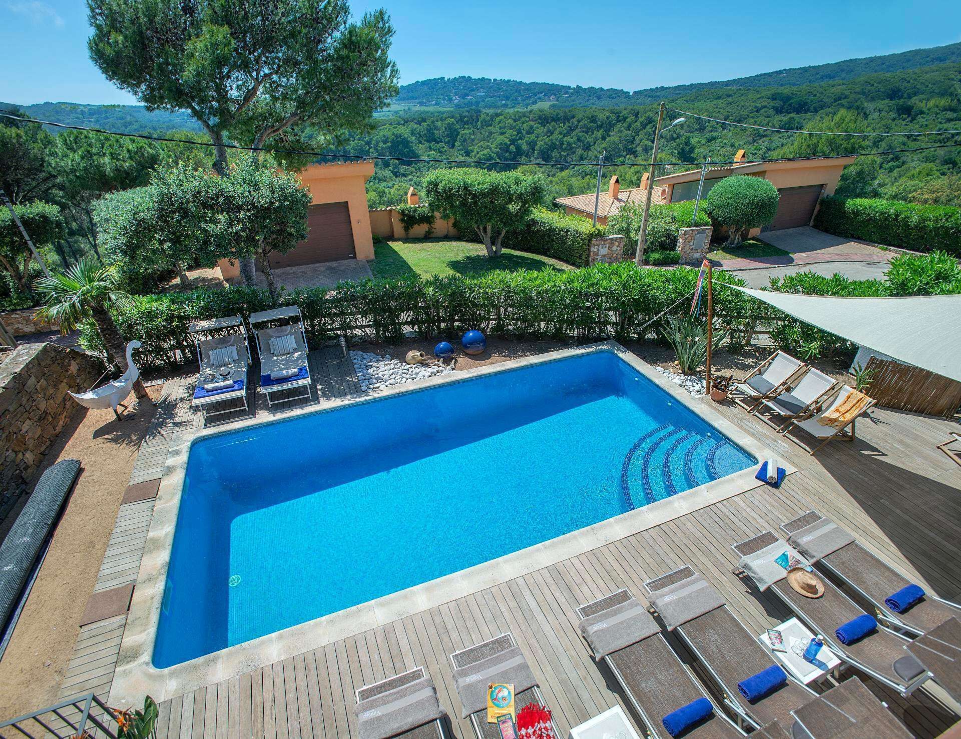 A rectangular swimming pool with a wooden deck and lounge chairs, surrounded by greenery and hills in the background.