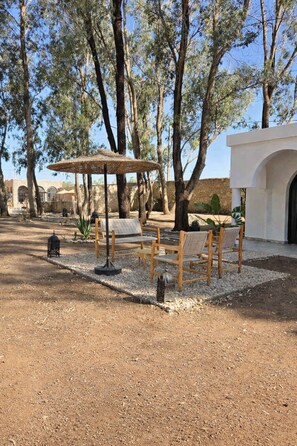 An outdoor seating area with wooden furniture and a straw canopy, surrounded by trees and a stone wall. - VILLA LATIFA Room in Agadir (Ghazoua)