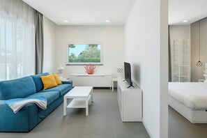 Quarto de hotel moderno com um sofá azul, uma escrivaninha branca, uma cama e uma janela com vista para vegetação.