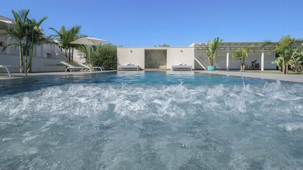 A swimming pool with water jets, palm trees, and a building with a white facade. - Modern villa with pool in Fontane Bianche (Siracusa)