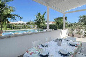 Área de refeições ao ar livre coberta, com mesa posta para quatro pessoas, com vista para uma piscina e vegetação exuberante.