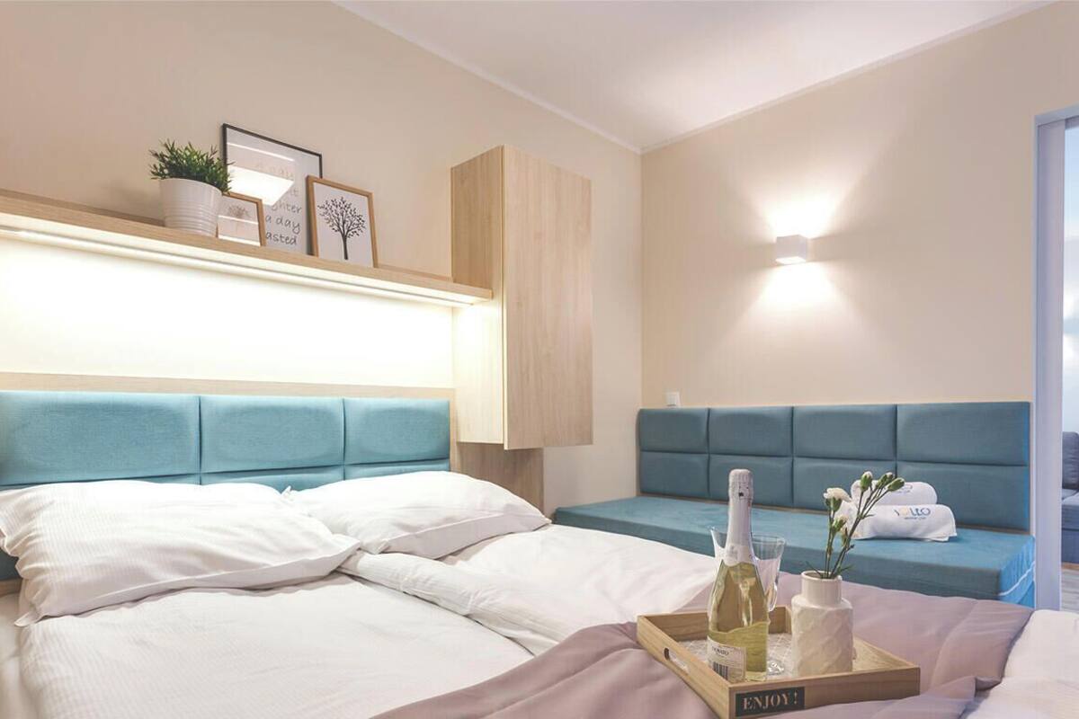 Habitación de hotel con cama, zona de estar, mesita de noche con una botella y flores, y una lámpara de pared.