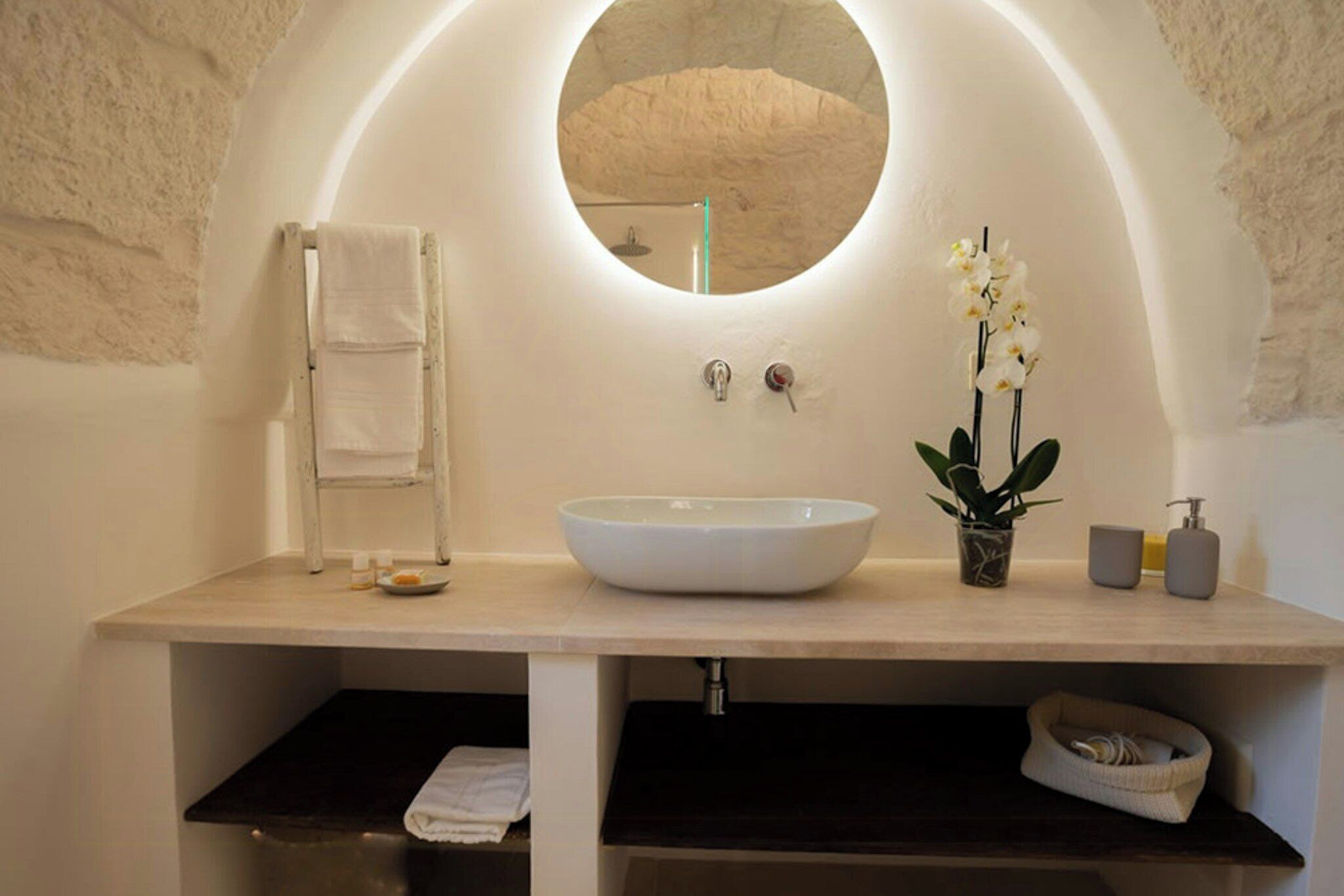 A bathroom with a round mirror, a white sink, a towel rack, a potted plant, and a soap dispenser.