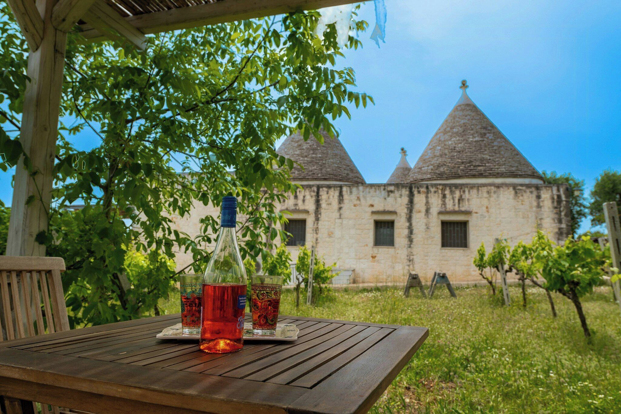 Ein Holztisch mit einer Flasche und Gläsern, eine Pergola und ein Steinhaus mit Kegeldächern im Hintergrund.