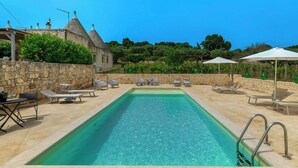 A rectangular pool with clear water, surrounded by lounge chairs and umbrellas, stone walls, and a building with conical roofs. - Apulian trullo with pool among vineyards (Martina Franca)