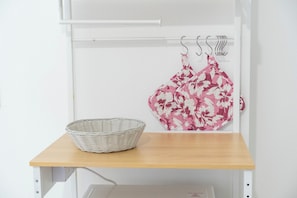 Eine weiße Wand mit einem weißen Regal, auf dem ein Korbwägelchen und ein pinkes Kleid mit Blumenmuster liegen.