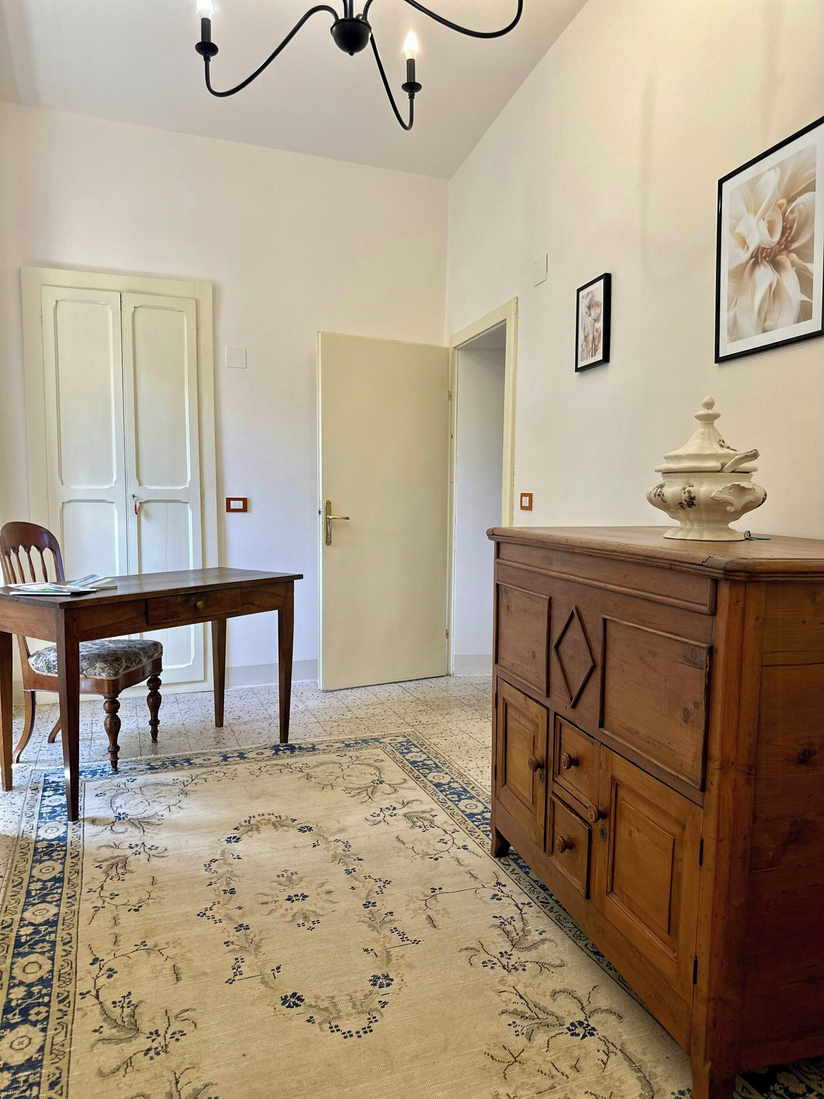 A room with a dining table, chairs, a wooden cabinet, a chandelier, and a patterned rug.
