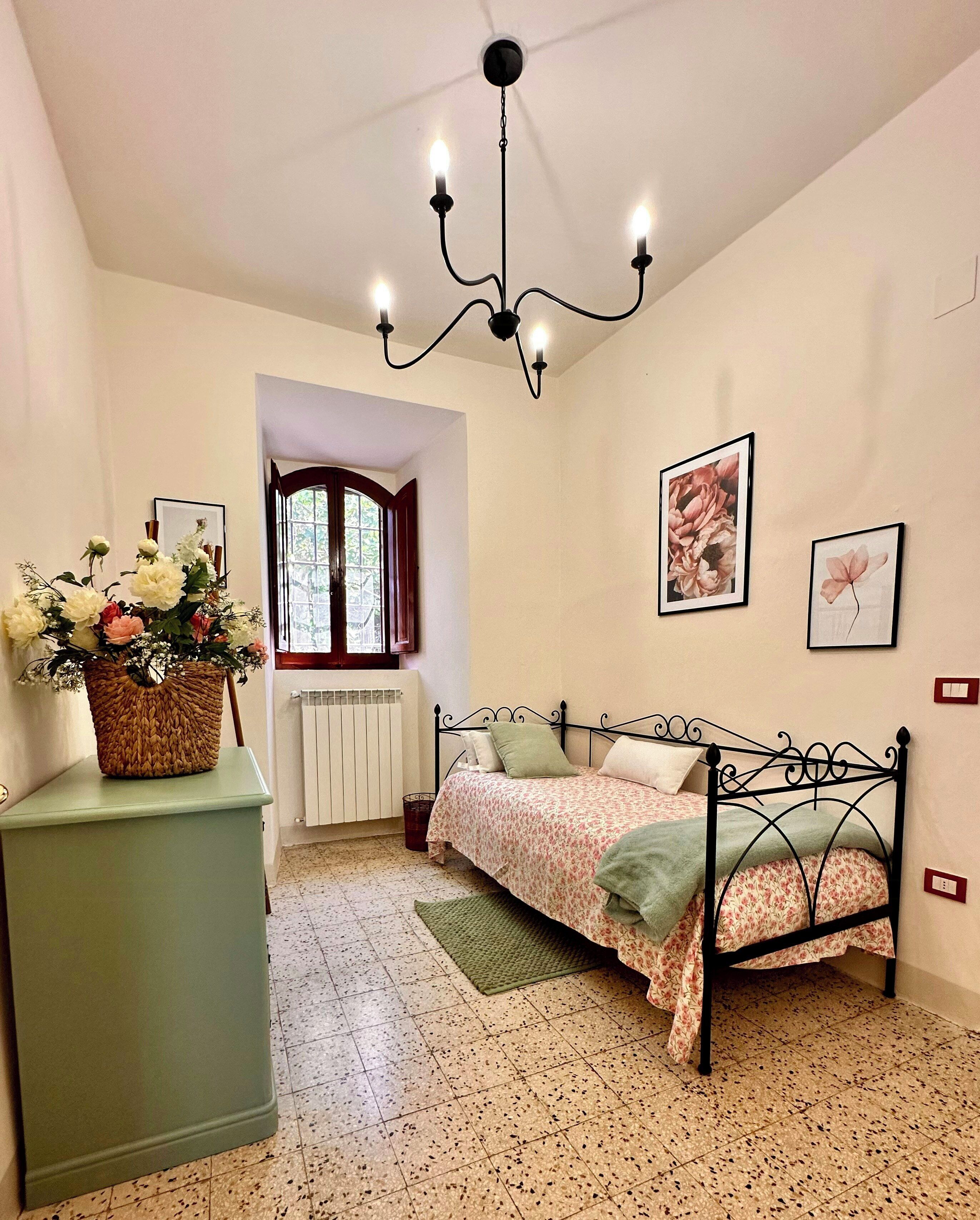 A bedroom with a bed, a radiator, a window, a chandelier, and a wall with framed pictures.