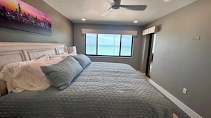 A bedroom with a large bed, a ceiling fan, a window with a view of the ocean, and a painting on the wall. - Riviera Club 1103 - Direct Beach Access (Fort Myers Beach)