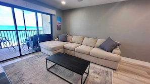 A living room with a sectional sofa, a coffee table, and a view of the ocean through large glass doors. - Riviera Club 1103 - Direct Beach Access (Fort Myers Beach)