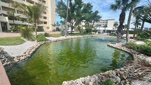 A landscaped pool area with a curved edge, surrounded by rocks and plants, and a multi-story building in the background. - Riviera Club 1103 - Direct Beach Access (Fort Myers Beach)
