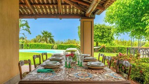 Un espace de restauration en plein air avec une table dressée pour six personnes, entourée de chaises, sous une pergola en bois offrant une vue sur une piscine et une végétation luxuriante.