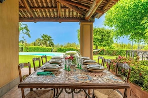 An outdoor dining area with a table set for six, surrounded by chairs, under a wooden pergola with a view of a swimming pool and lush greenery.