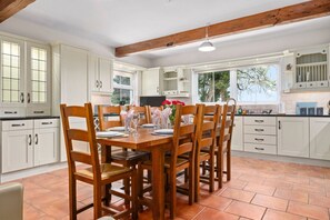 A kitchen with white cabinets, a wooden dining table, and chairs. There are windows and a view of trees outside. - Hilltop View by Escape Ordinary (Lisbellaw, Enniskillen)