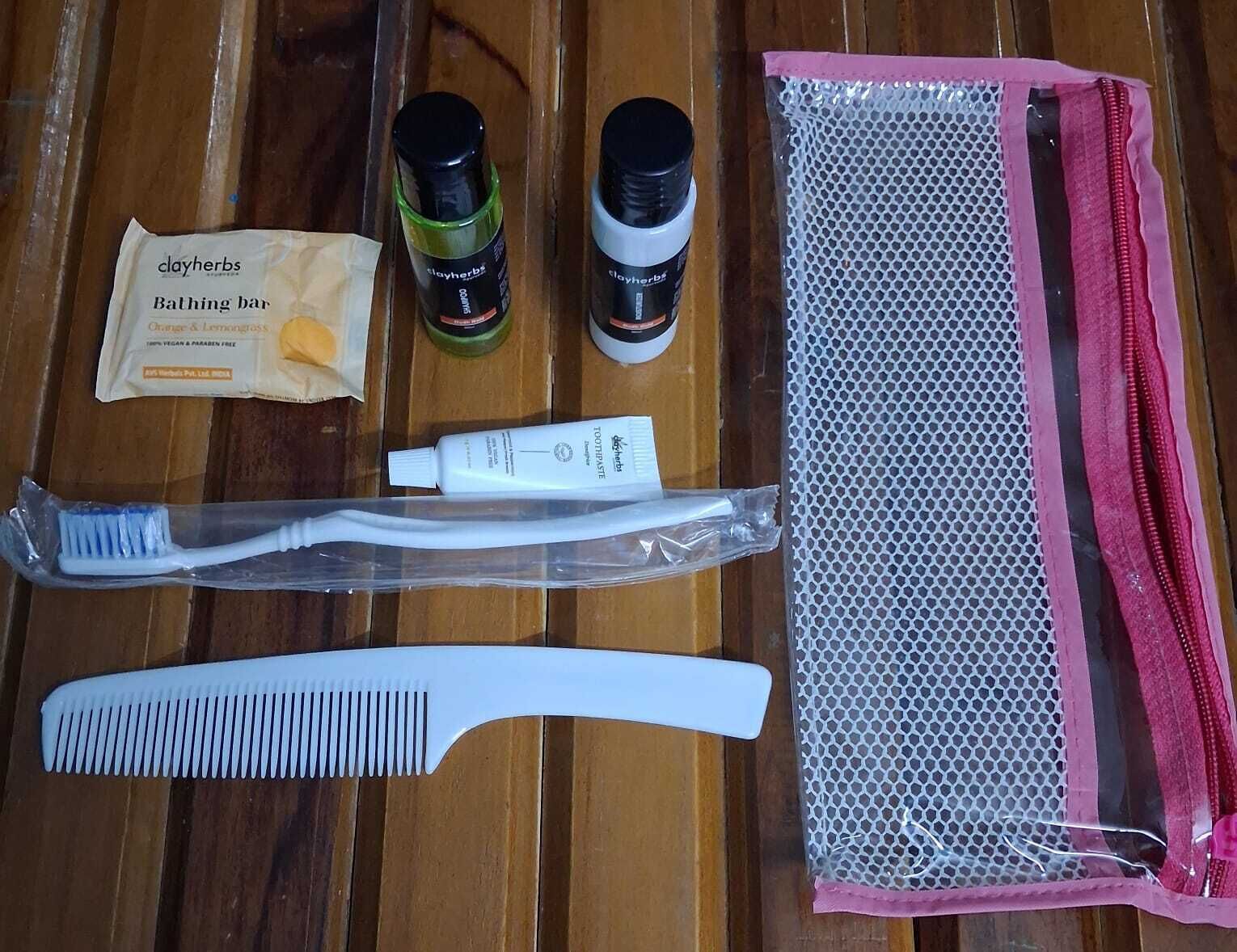 Una superficie de madera con un paquete etiquetado como 'clayherbs Jabón de baño Naranja y Hierba Limón', dos botellas de 'clayherbs', un tubo de 'clayherbs Pasta dental' y un peine blanco, junto con una bolsa de malla.