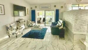 A spacious living room with a marble floor, a blue rug, a chandelier, and a large window with curtains. - Waterfront 3BR 1Bath Furnished w/Gulf of America Access (NEW PORT RICHEY)