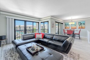 A modern living room with a sectional sofa, a dining area with red chairs, and large windows offering a view of buildings and greenery. - ☀️🏖️Gorgeous Bay View | Steps to the Beach | Santa Maria #205- Manatee Cove (Fort Myers Beach)