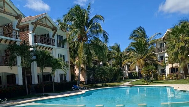 A clear blue pool surrounded by palm trees and a resort building with balconies. - Apartamento playa, piscina vabaro (Punta Cana)