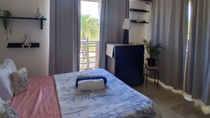 A bedroom with a bed, a desk, a chair, a shelf with decorative items, and a window with curtains. - Apartamento playa, piscina vabaro (Punta Cana)