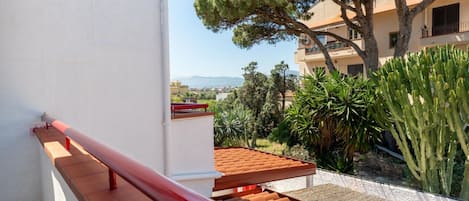 A balcony with a red railing, a tiled roof, and a view of a building with a tree and cacti.