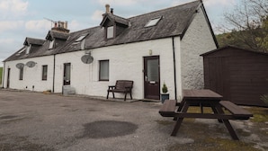 A white building with multiple windows, a door, and a chimney. There is a bench and a picnic table outside. - 3 Bruaich Cottages (Strathpeffer)