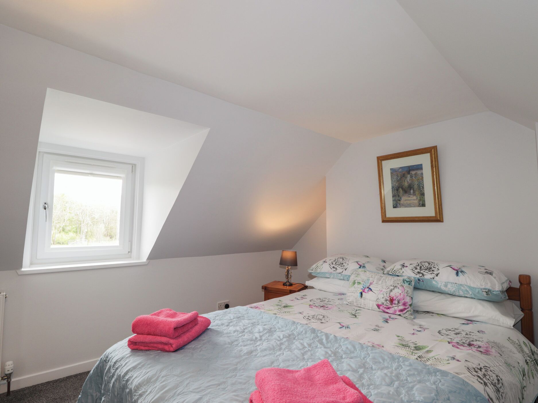 A bedroom with a sloped ceiling, a window, a bed with floral bedding, a bedside lamp, and a framed picture on the wall.