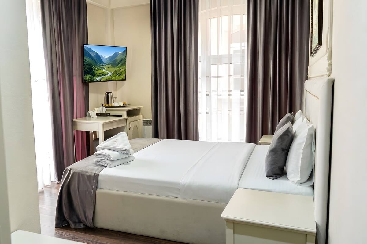 A hotel room with a bed, bedside tables, a desk with a flat-screen TV, and curtains.