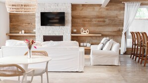 Una sala de estar moderna con pared de piedra, vigas de madera, televisor de pantalla plana, mobiliario blanco y un comedor.