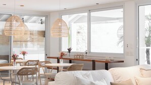 Un interior moderno con sofá blanco, mesa de comedor, lámparas colgantes de mimbre y ventanas grandes.