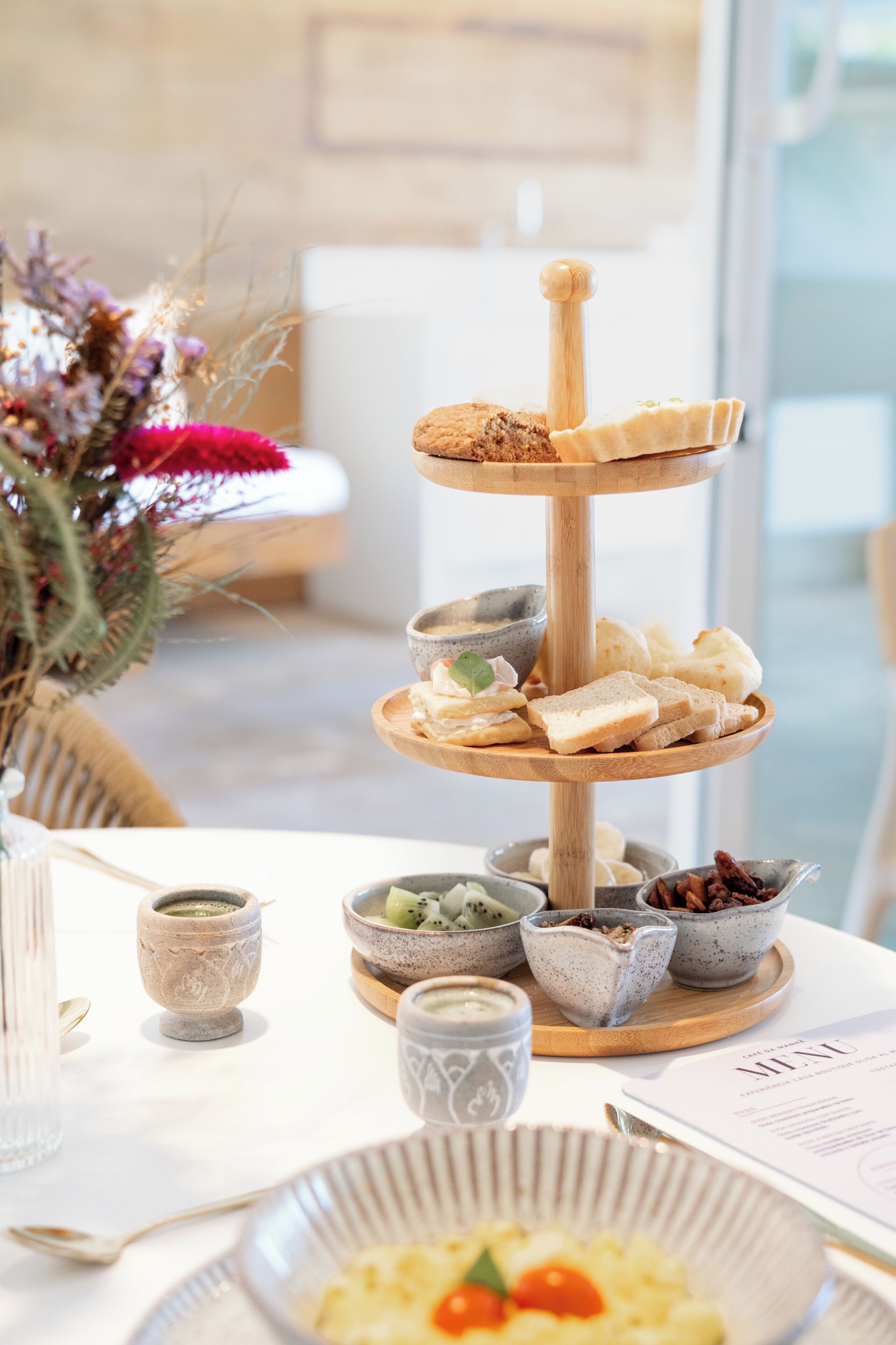 A three-tiered wooden stand holding various food items, a vase with dried flowers, and a menu on the table.