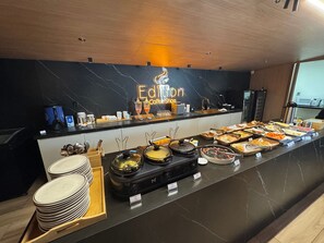 Een buffet met een marmeren achterwand, een neonbord waarop 'Edition Coffee Shop' staat, en diverse etenswaren en bestek.